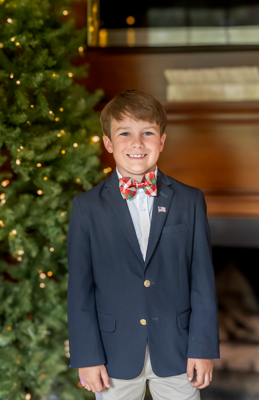 Young boy in a navy blazer with a festive bow tie standing in front of a decorated Christmas tree.